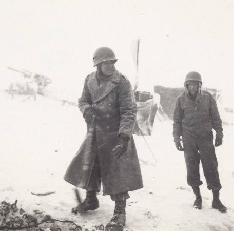 Luxembourg, December 1944: Members of the 180th Field Artillery Battalion, 26th Infantry Division try to keep warm during a driving snowstorm.  The Soldier in foreground wears his Melton wool overcoat, while throwing wood into the fire. Note the Soldier in the background reliant upon the inadequate Olive Drab Field Jacket.
