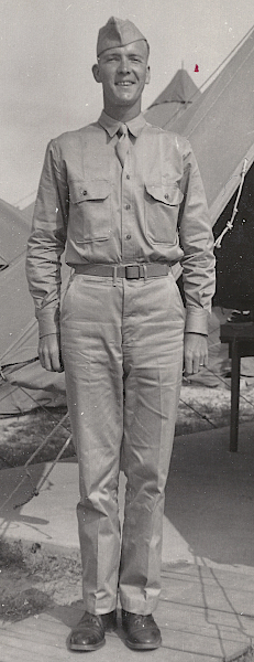 Pvt. Hogan wears the summer service uniform featuring the khaki cotton field cap and necktie. The shirt is specification QMC 6‐241 with standing and falling collar and front button placket.  Below the belt on his right side is a watch pocket with double welt construction indicating his trousers are specification QMC 6‐254.  Note the olive‐drab web belt with frame buckle.